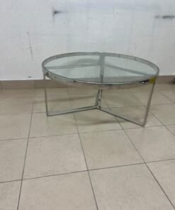 A round coffee table featuring a silver-colored metal frame (steel or chrome), a clear glass top, and a geometric crisscross base design.