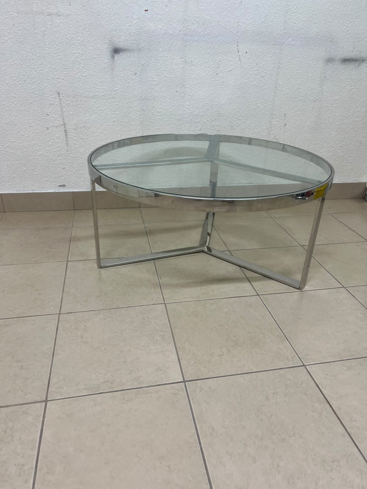 A round coffee table featuring a silver-colored metal frame (steel or chrome), a clear glass top, and a geometric crisscross base design.