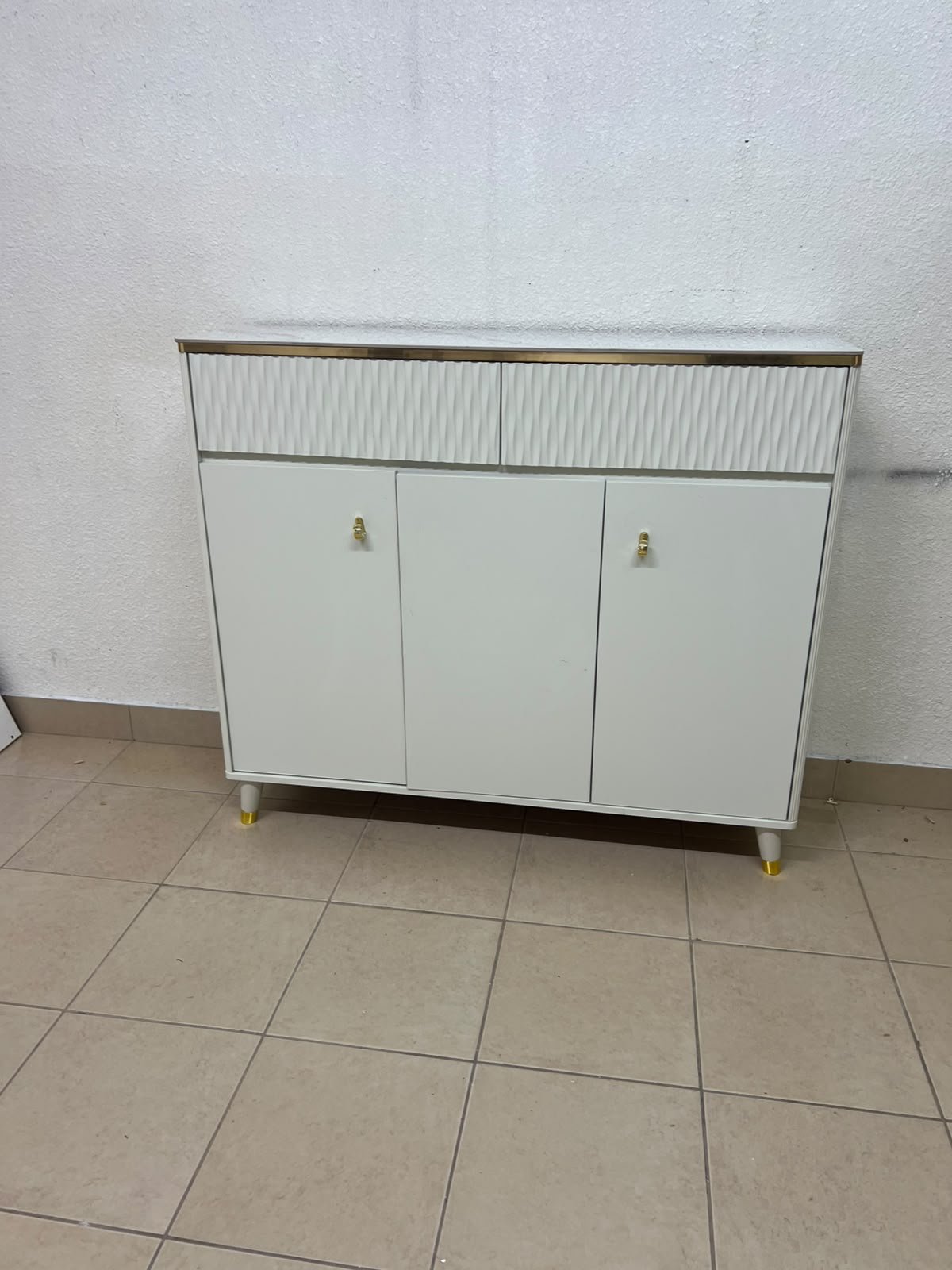 A white shoe cabinet or entryway storage unit (console) featuring a modern design, ribbed upper drawers, and gold accents on the handles and legs.