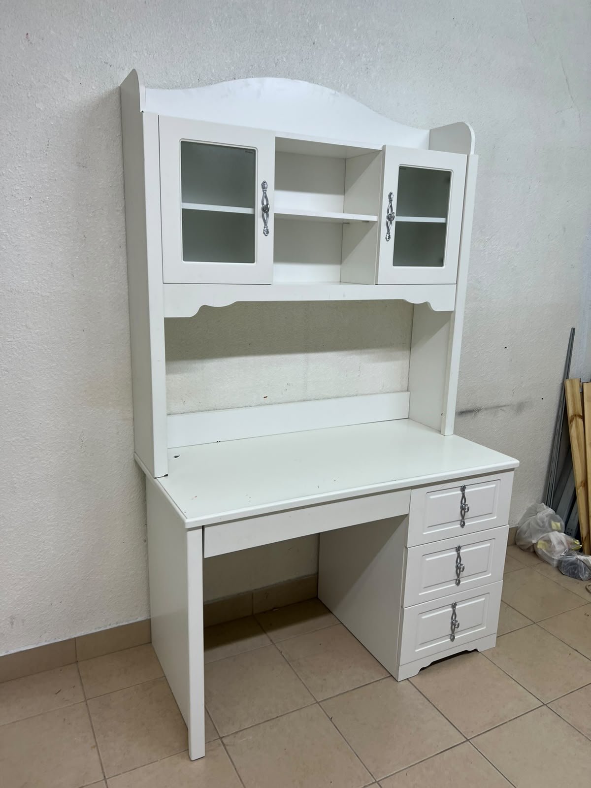 A complete white study desk featuring an overhead hutch, distinguished by its upper cabinets with glass doors and decorative handles.