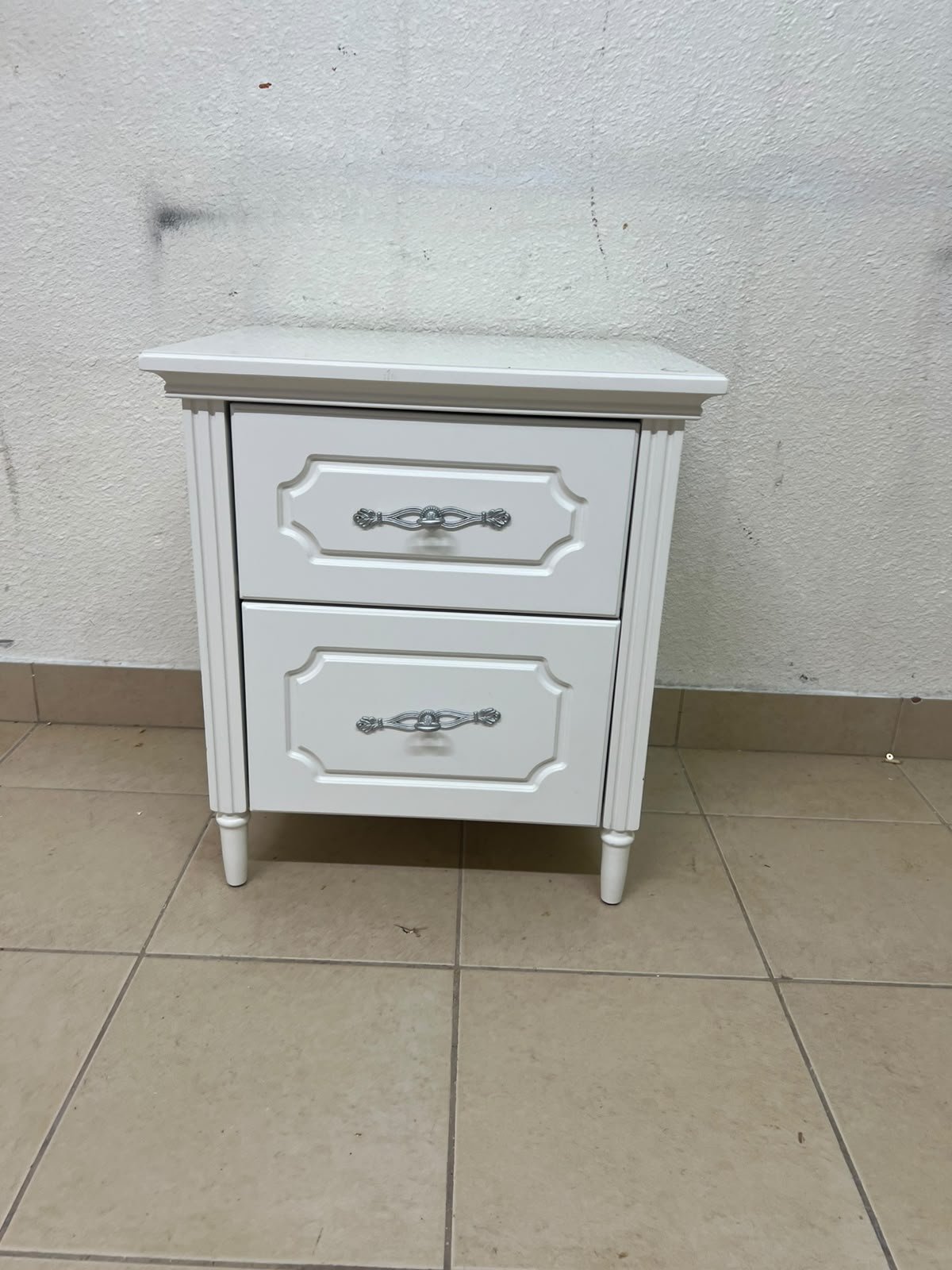 The elegant white side table with two drawers is distinguished by its decorative details and prominent silver handles.