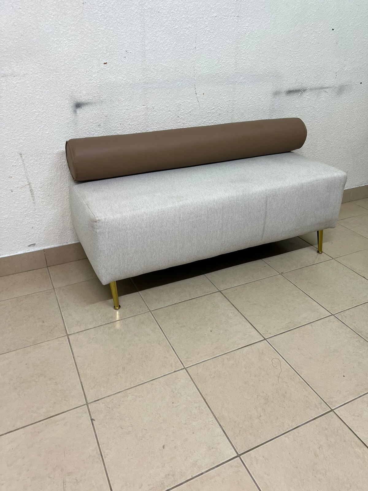 The light-gray upholstered seat, featuring a brown cylindrical backrest and gold legs, is a stylish, modern piece of furniture that adds a distinctive touch to the décor.