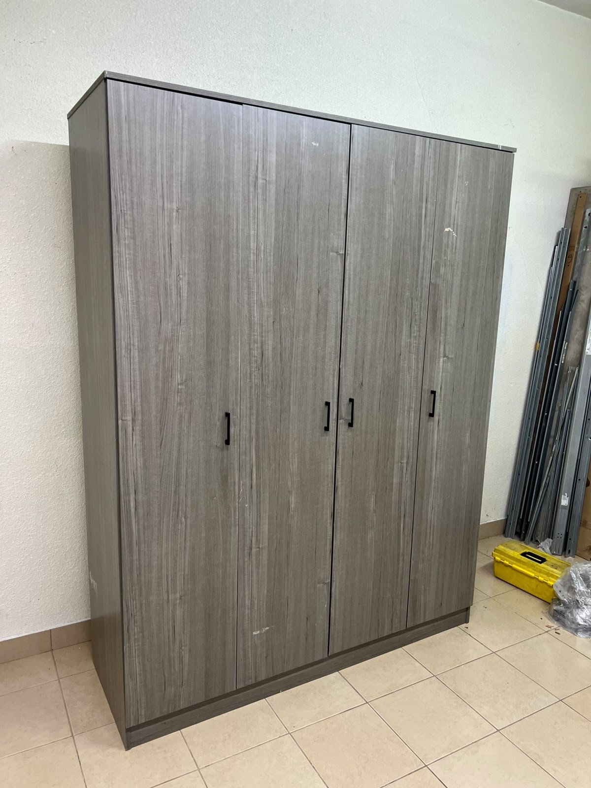 The image shows the Jamora four-door wardrobe from Danube Home, featuring a warm grey color and a practical design with hinged doors and simple black handles.