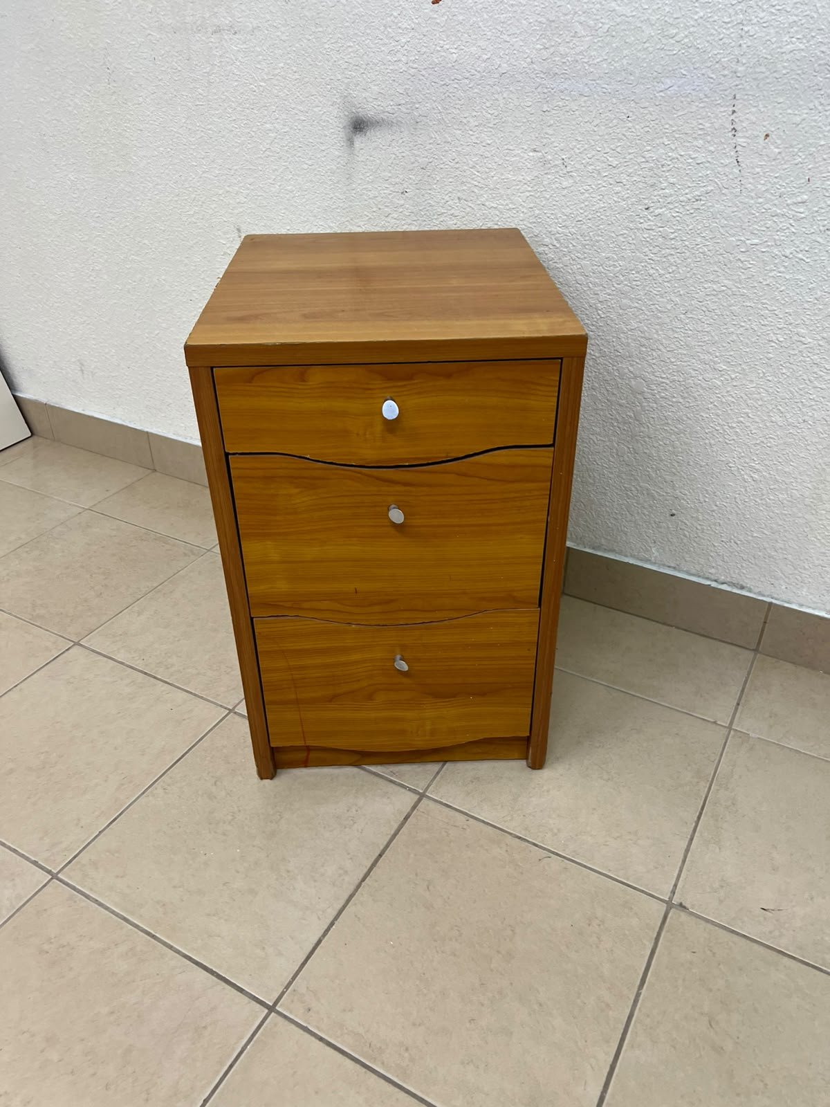 A wooden side table (nightstand) featuring three drawers with slightly curved fronts and small, round handles.