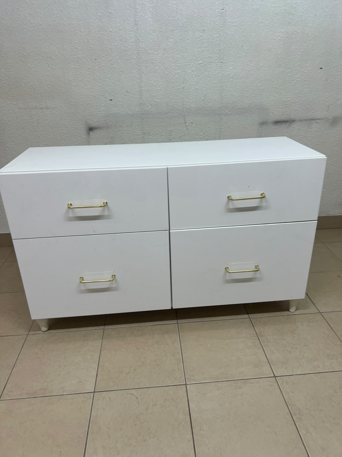 A modern, low-profile white chest of drawers (dresser) featuring a soft white finish, four spacious drawers (or double-fronted drawers), and gold or copper-toned pulls, all set on raised white legs. The design resembles contemporary storage units like the IKEA BESTÅ system or similar four-drawer cabinets.