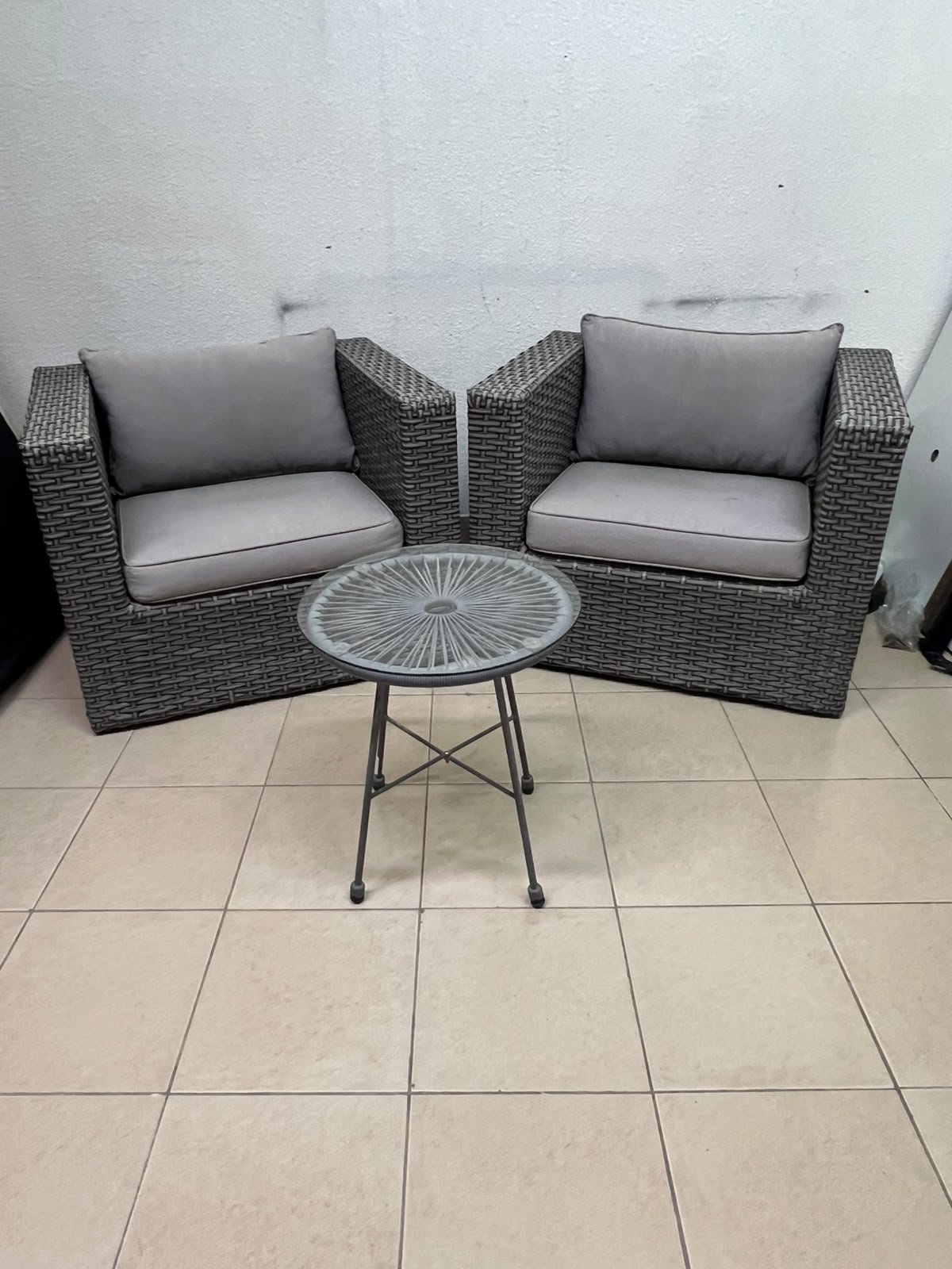 A compact outdoor seating set consisting of two grey synthetic rattan (PE Rattan) armchairs with comfortable grey cushions, plus a round table with a glass top.