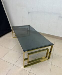 A modern coffee table featuring a dark glass top (tempered or smoked glass) and a shiny gold metal frame and base, distinguished by an interlocking geometric design on the sides. - Image 2