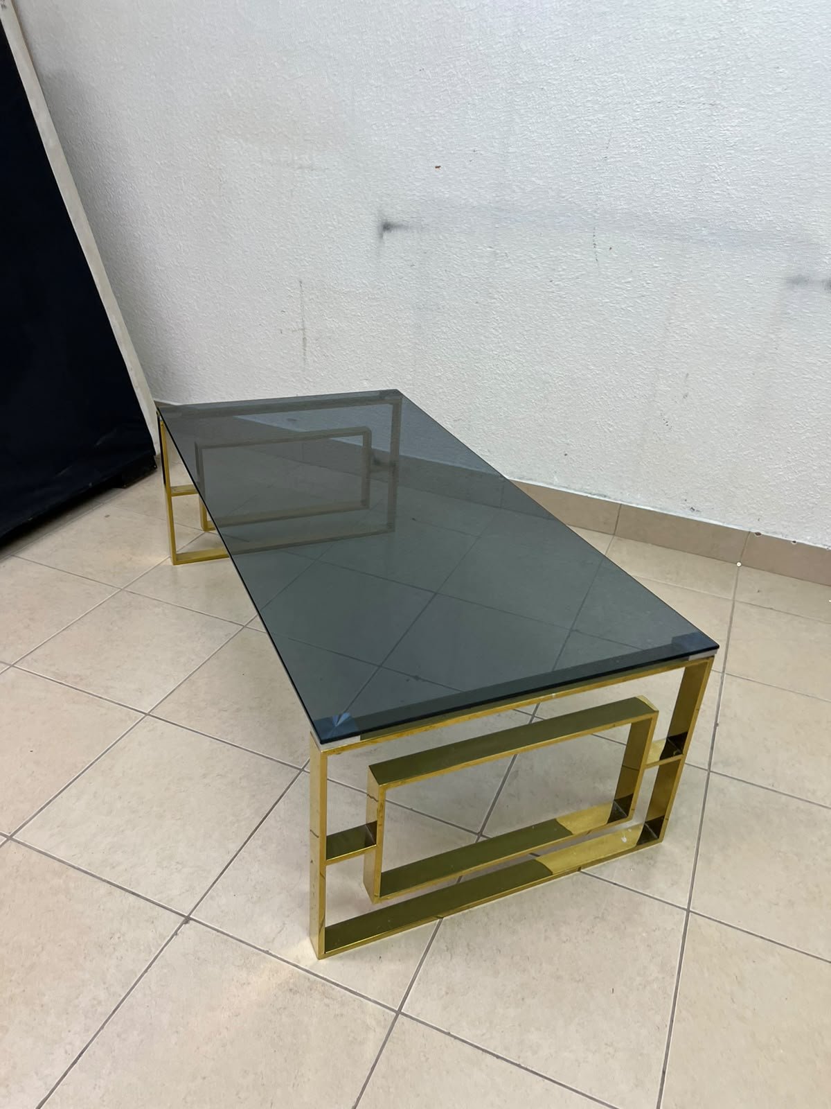 A modern coffee table featuring a dark glass top (tempered or smoked glass) and a shiny gold metal frame and base, distinguished by an interlocking geometric design on the sides. - Image 2