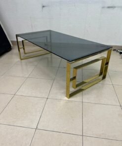 A modern coffee table featuring a dark glass top (tempered or smoked glass) and a shiny gold metal frame and base, distinguished by an interlocking geometric design on the sides.