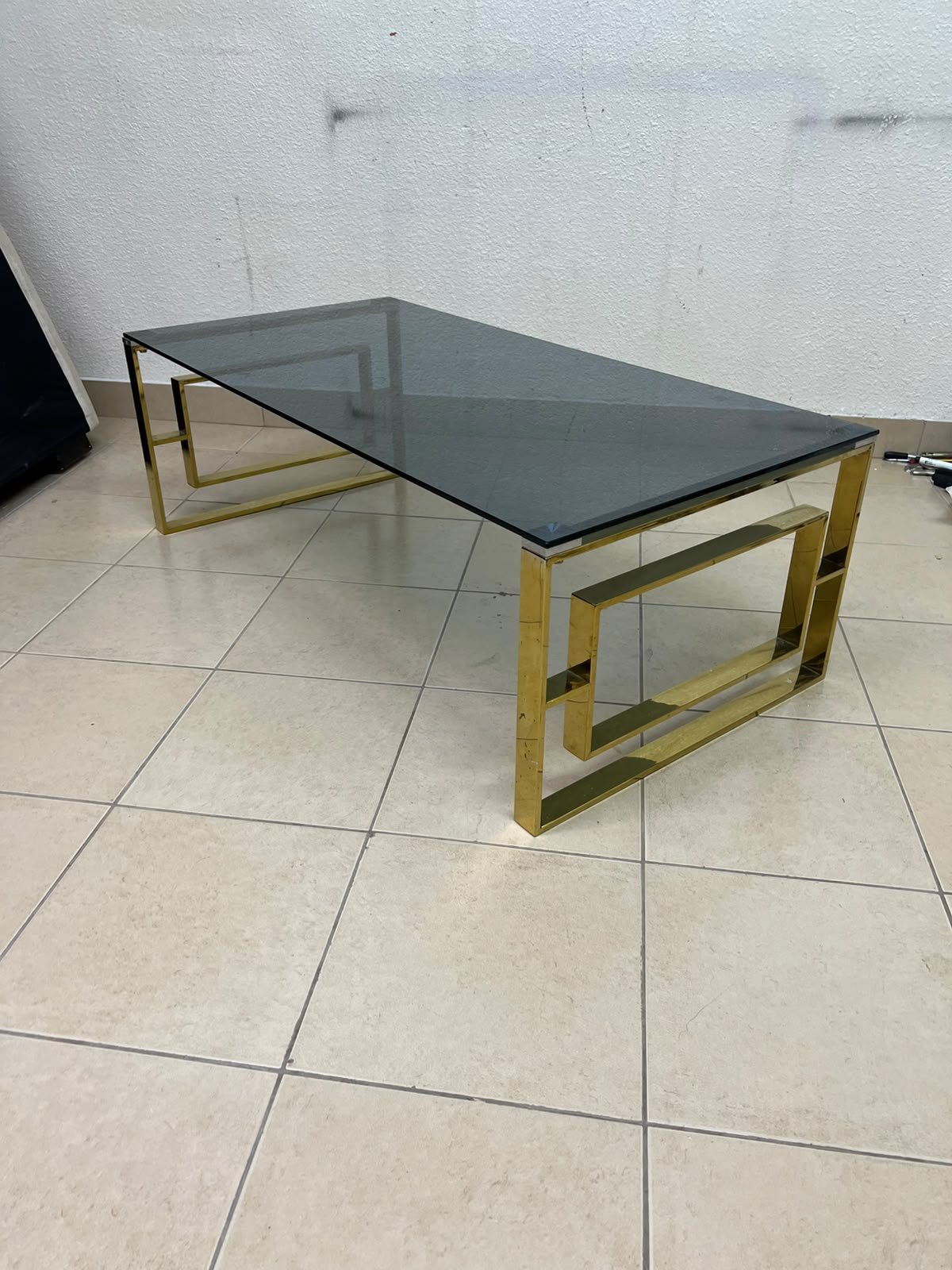 A modern coffee table featuring a dark glass top (tempered or smoked glass) and a shiny gold metal frame and base, distinguished by an interlocking geometric design on the sides.