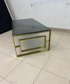 A modern coffee table featuring a dark glass top (tempered or smoked glass) and a shiny gold metal frame and base, distinguished by an interlocking geometric design on the sides. - Image 4
