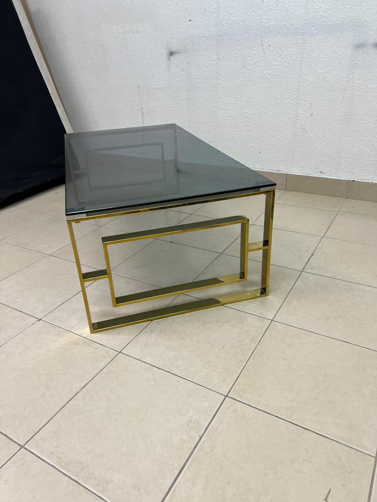 A modern coffee table featuring a dark glass top (tempered or smoked glass) and a shiny gold metal frame and base, distinguished by an interlocking geometric design on the sides. - Image 4
