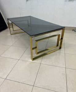 A modern coffee table featuring a dark glass top (tempered or smoked glass) and a shiny gold metal frame and base, distinguished by an interlocking geometric design on the sides. - Image 5