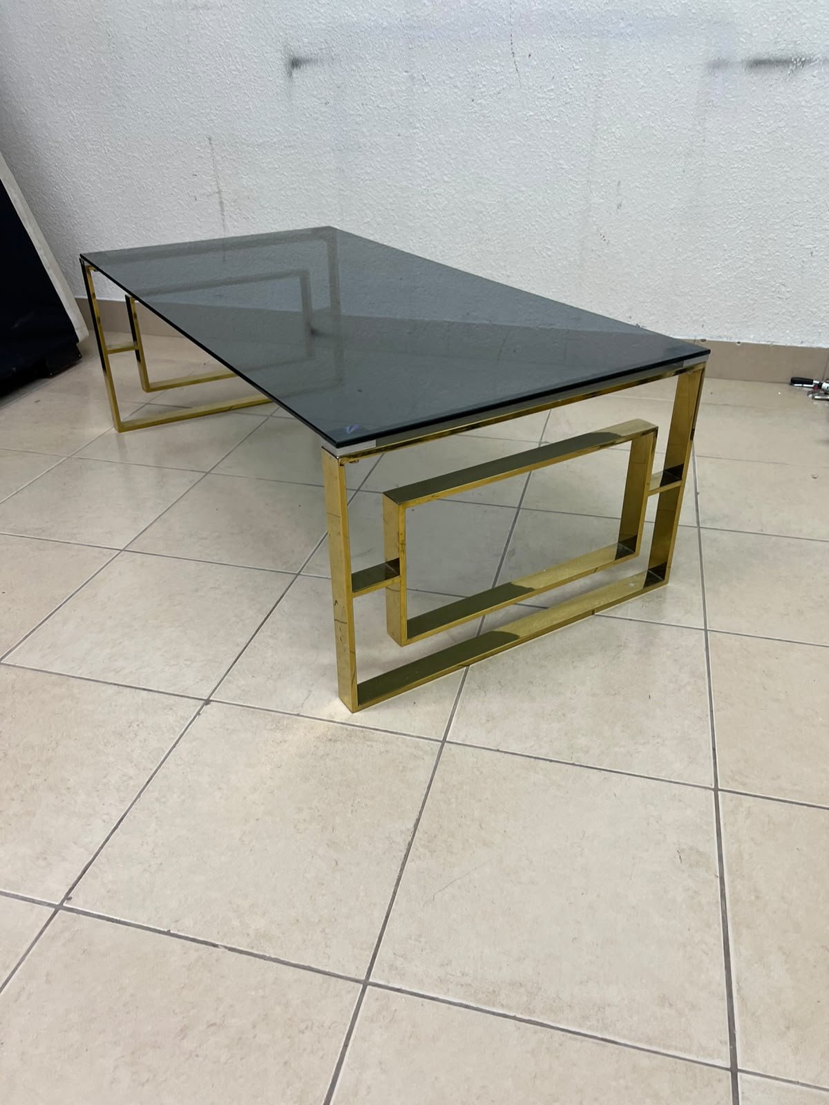 A modern coffee table featuring a dark glass top (tempered or smoked glass) and a shiny gold metal frame and base, distinguished by an interlocking geometric design on the sides. - Image 5