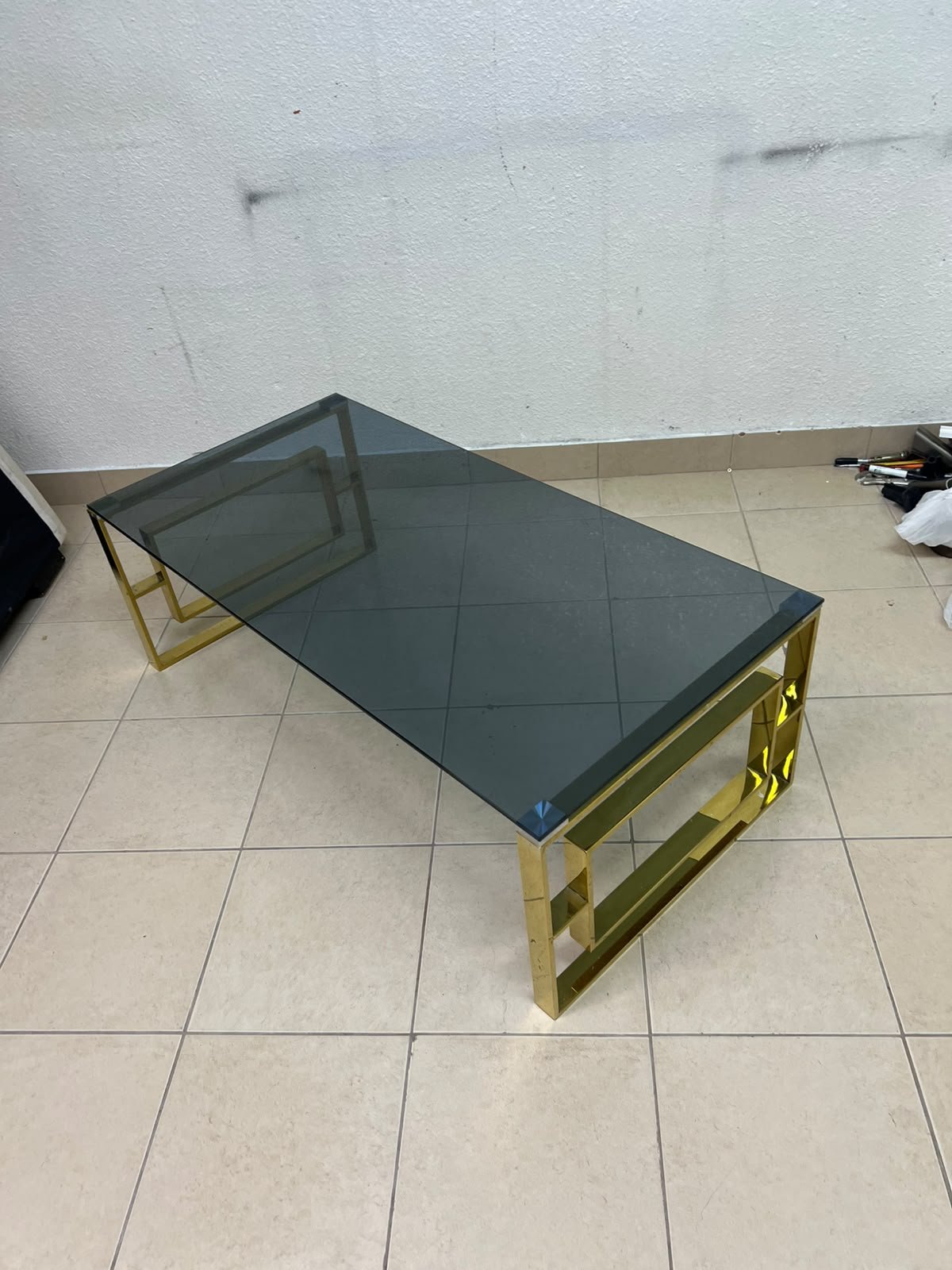 A modern coffee table featuring a dark glass top (tempered or smoked glass) and a shiny gold metal frame and base, distinguished by an interlocking geometric design on the sides. - Image 9