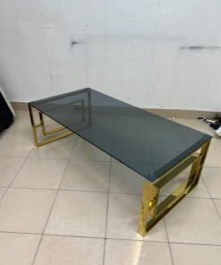 A modern coffee table featuring a dark glass top (tempered or smoked glass) and a shiny gold metal frame and base, distinguished by an interlocking geometric design on the sides. - Image 8