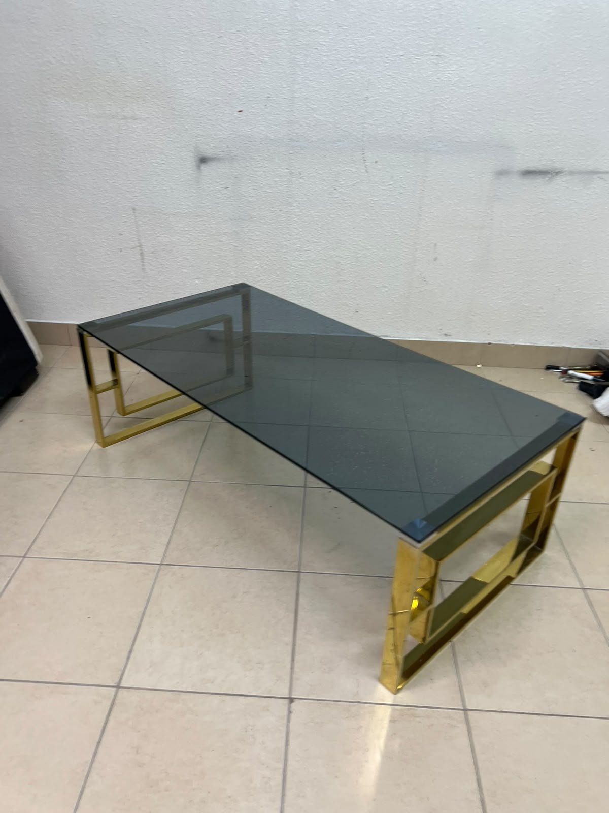 A modern coffee table featuring a dark glass top (tempered or smoked glass) and a shiny gold metal frame and base, distinguished by an interlocking geometric design on the sides. - Image 8
