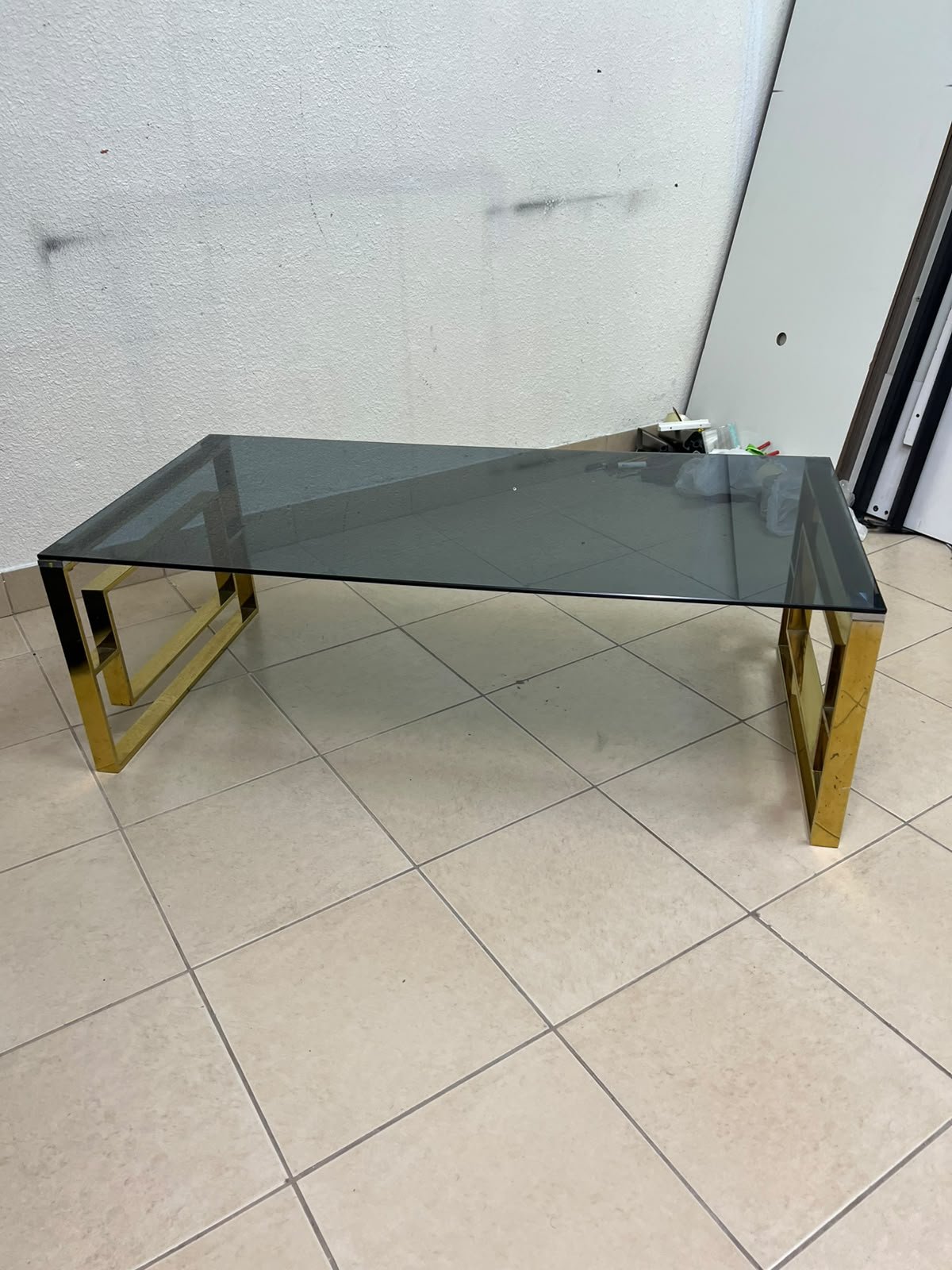 A modern coffee table featuring a dark glass top (tempered or smoked glass) and a shiny gold metal frame and base, distinguished by an interlocking geometric design on the sides. - Image 7