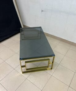 A modern coffee table featuring a dark glass top (tempered or smoked glass) and a shiny gold metal frame and base, distinguished by an interlocking geometric design on the sides. - Image 6