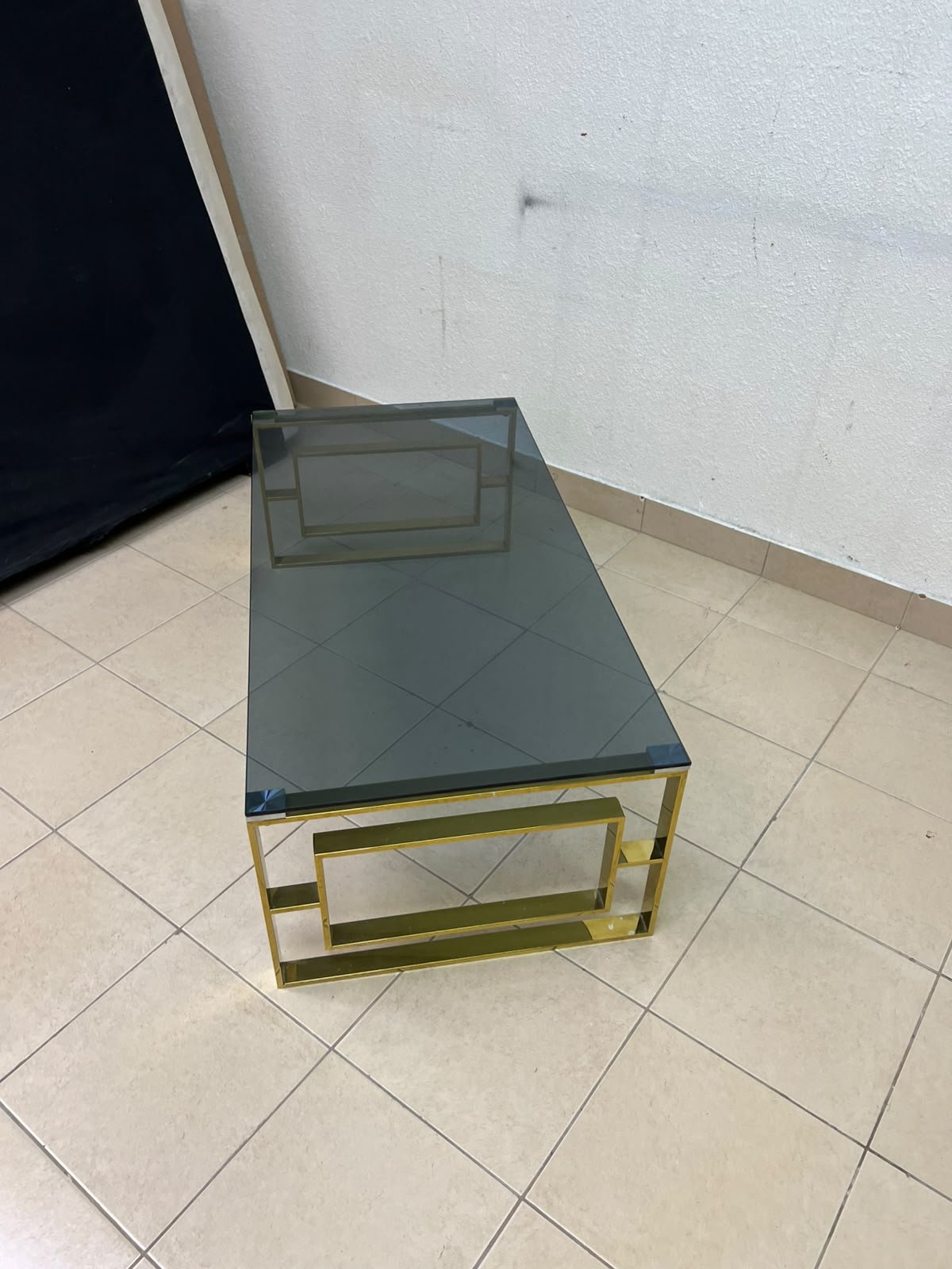 A modern coffee table featuring a dark glass top (tempered or smoked glass) and a shiny gold metal frame and base, distinguished by an interlocking geometric design on the sides. - Image 6