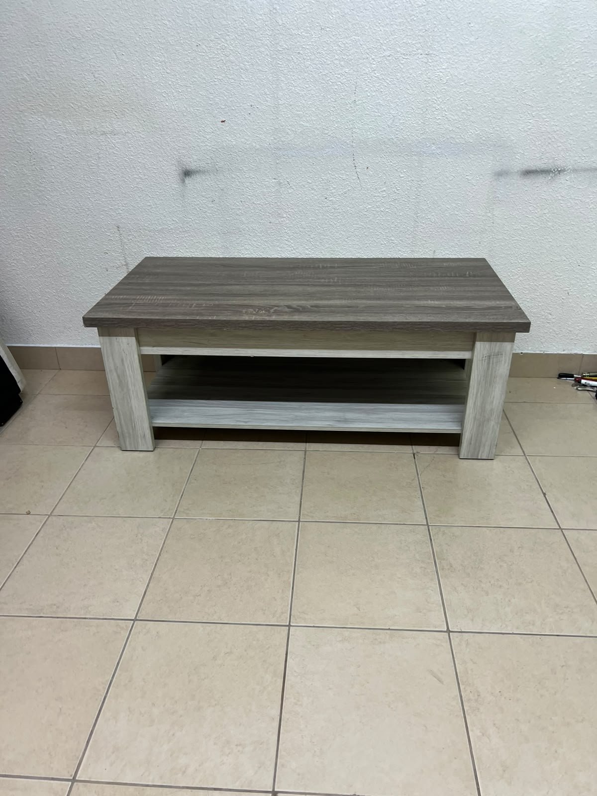 A rectangular coffee table made of engineered wood featuring a two-tone design; the tabletop comes in a greyish-brown wood finish, while the frame, legs, and lower shelf are white or light grey. It resembles the "Angelic" coffee table from Home Box.