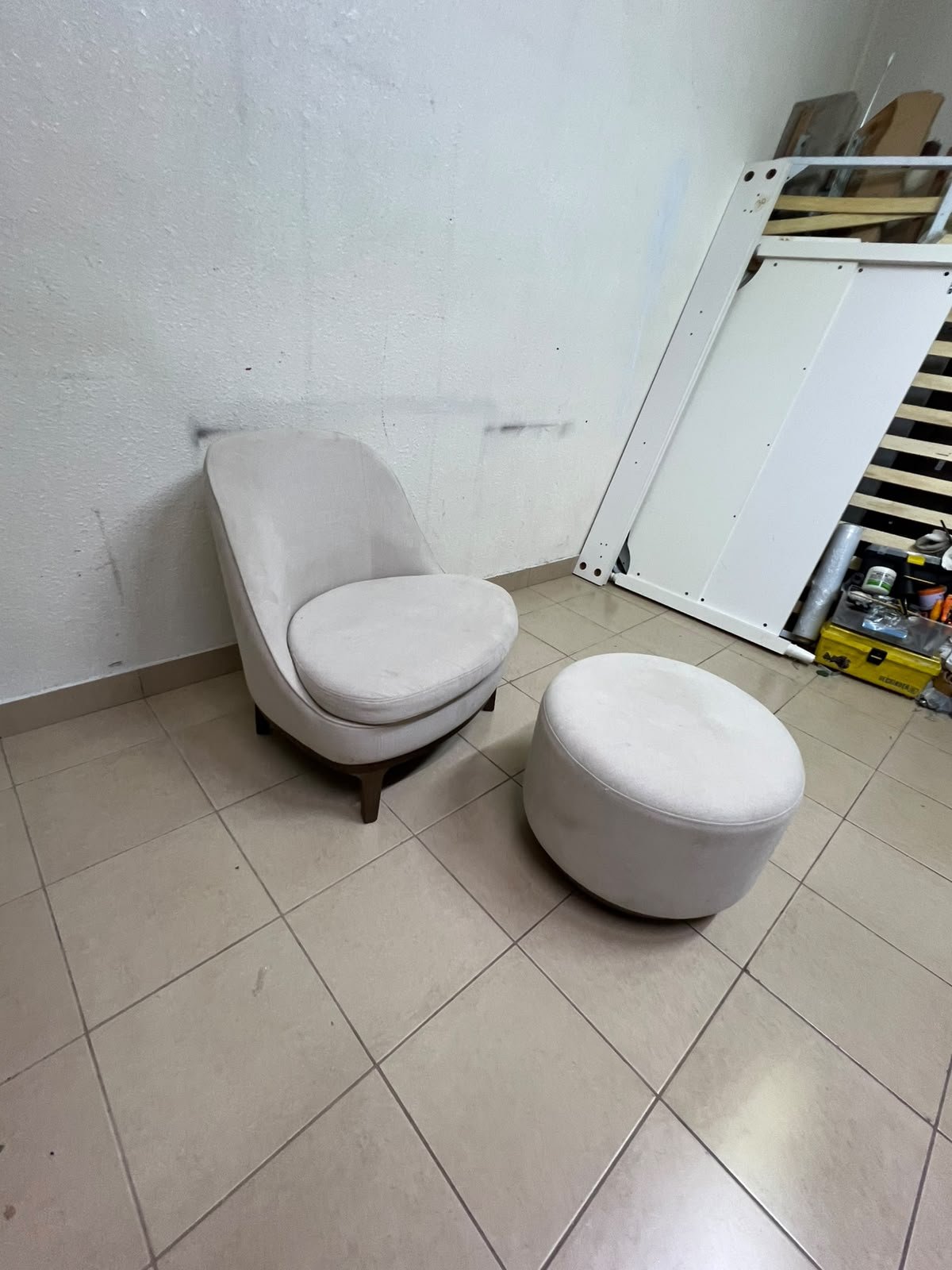 An armchair (poltrona) with a matching footstool (pouffe), featuring a modern, curved design; upholstered in a light beige velvet or linen-like fabric and set upon elegant wooden legs.