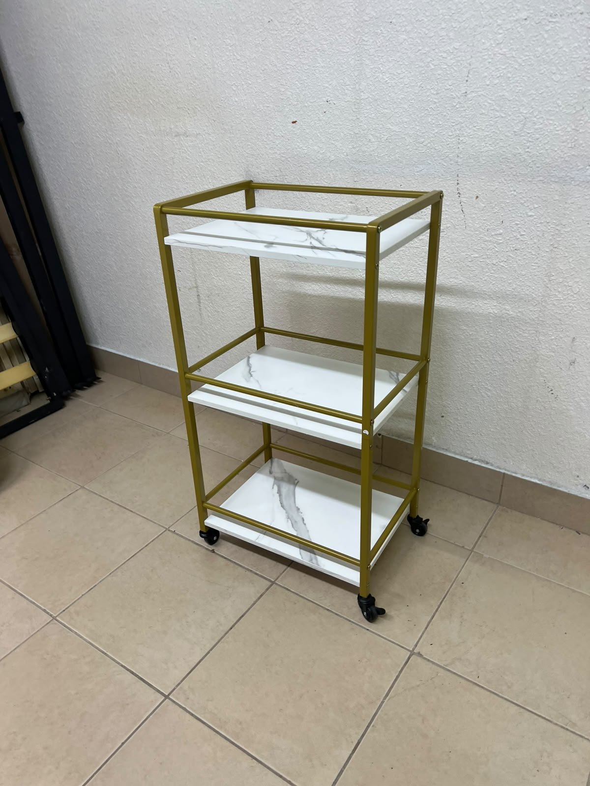 A 3-tier storage or serving cart on wheels, featuring a gold-colored metal frame and white, marble-design shelves.