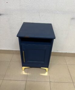 A bedside table (or small storage unit) in a deep blue (indigo) shade, featuring an open storage space at the top, a lower cabinet with a door fitted with a long, gold-colored handle, and elevated, gold-colored metal legs.