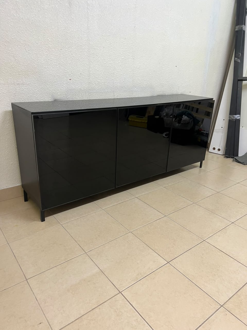 A cabinet (such as a TV stand or sideboard) that appears as a glossy black storage unit with three doors and slender legs; it bears a strong resemblance to the IKEA BESTÅ collection (specifically the Selsviken/Stubbarp combination in black-brown with high-gloss doors).