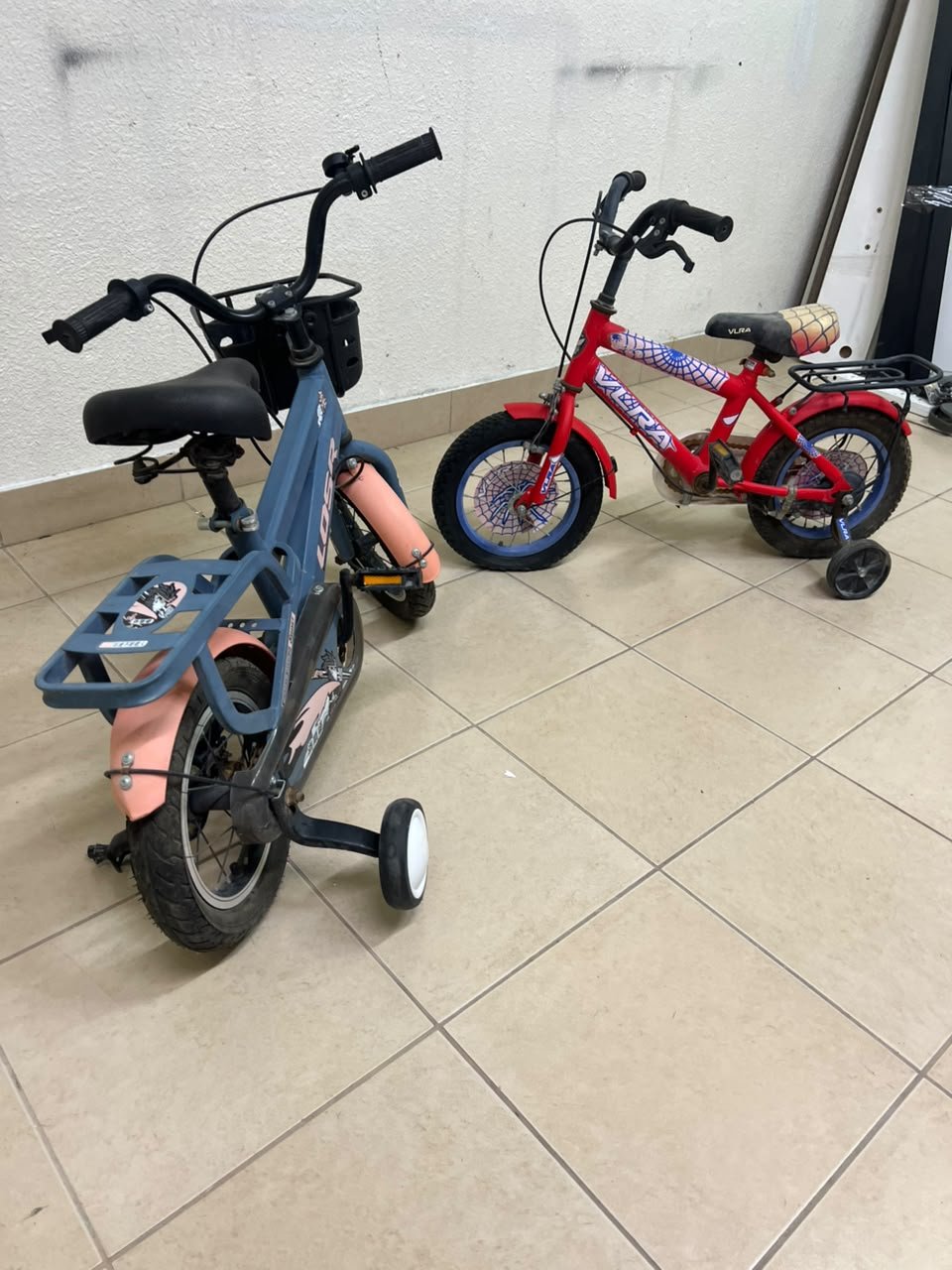 Two children’s bicycles in different colors (blue and red), equipped with training wheels and front baskets. - Image 6
