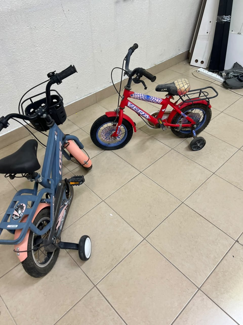 Two children’s bicycles in different colors (blue and red), equipped with training wheels and front baskets.