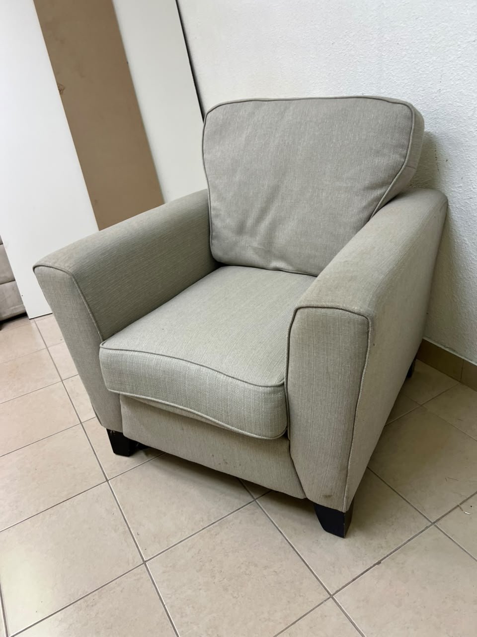 A single-seater fabric armchair in beige or light gray, featuring a separate seat cushion and back cushion, along with small black legs.
